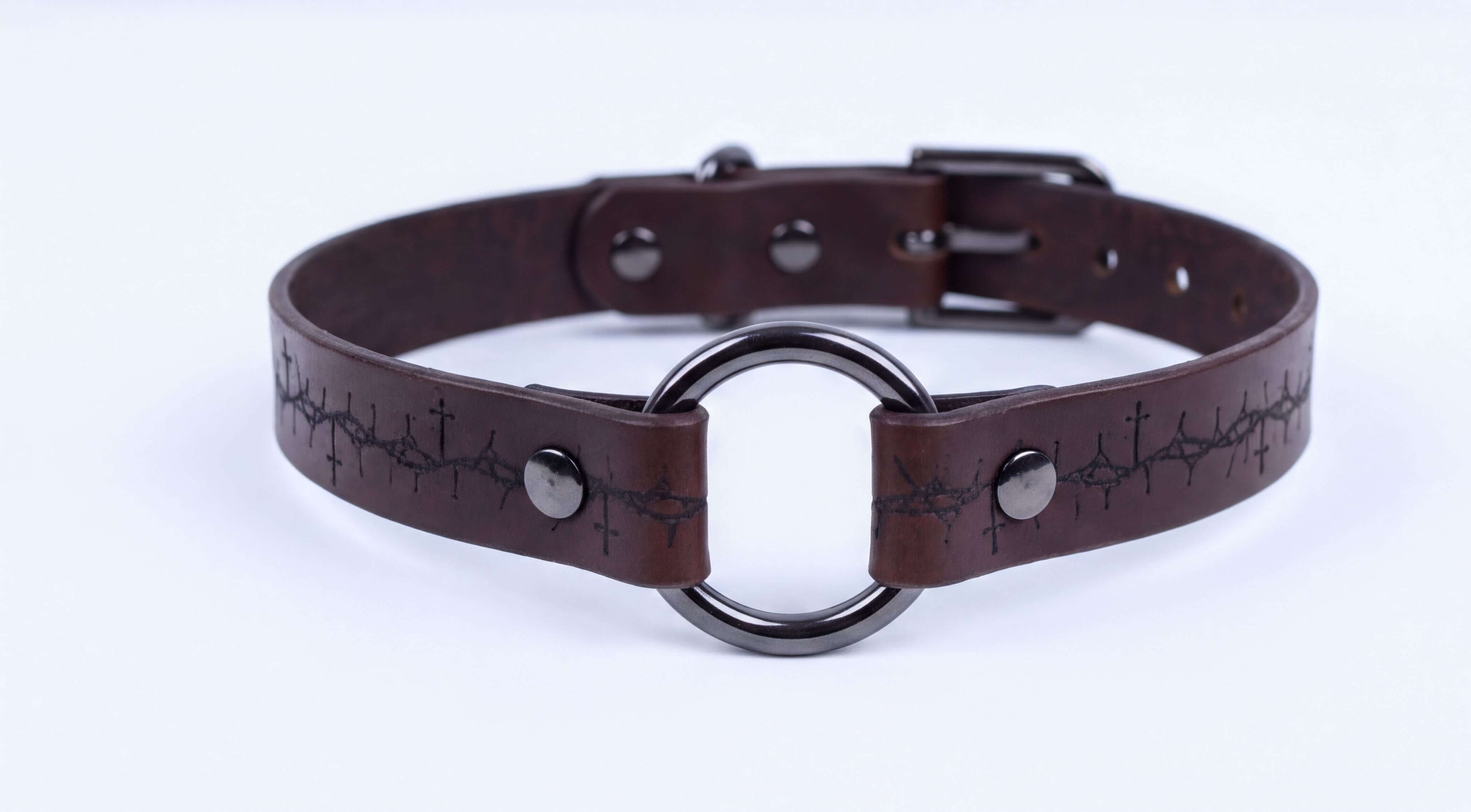 A brown leather choker collar with a metal ring and a cross pattern on the strap.