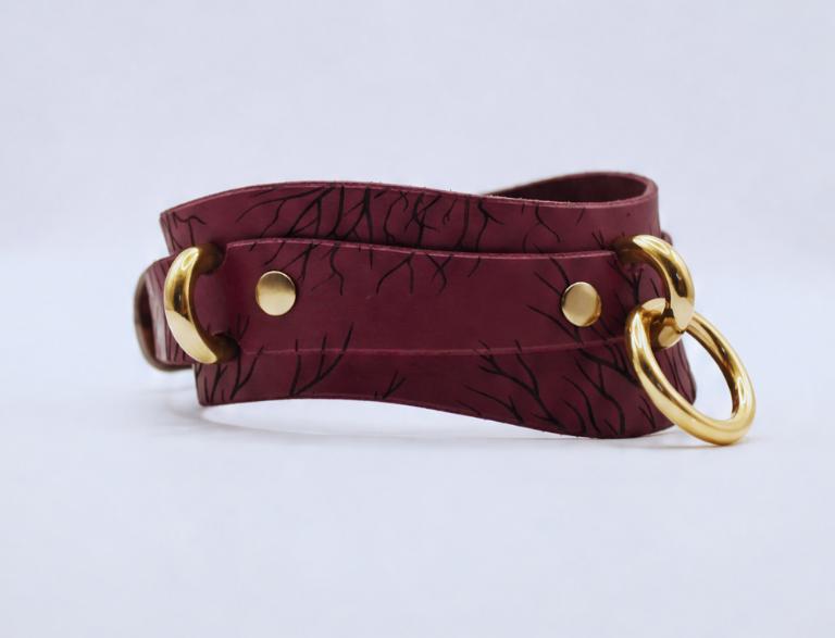 A burgundy leather choker necklace with a gold-colored metal ring and two gold-colored metal rings on either side.