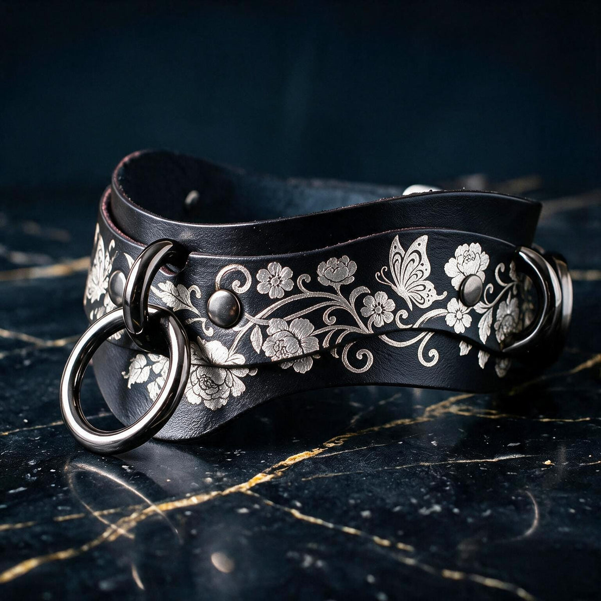A black leather strap with a floral pattern and metal rings on the ends, resting on a dark surface.