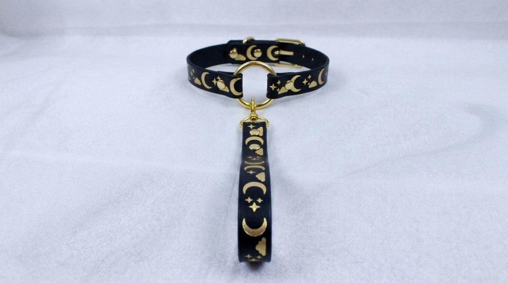 A black collar with gold moon and star designs, featuring a gold-colored loop and a dangling charm.