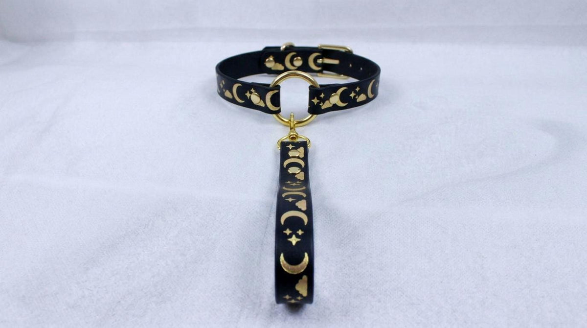 A black collar with gold moon and star designs, featuring a gold-colored loop and a dangling charm.