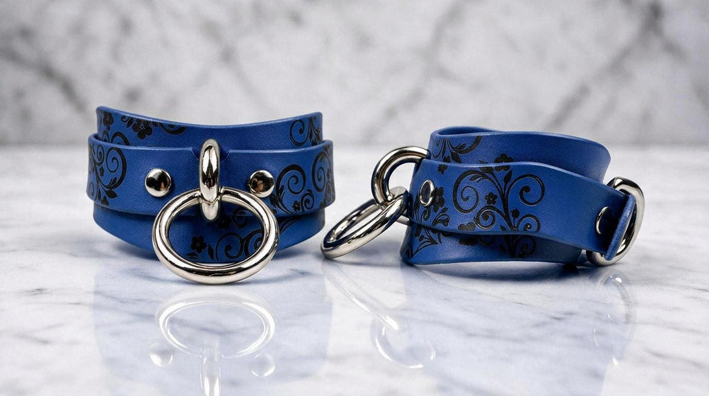 A pair of blue leather cuffs with silver metal rings and decorative patterns on them, placed on a marble surface.