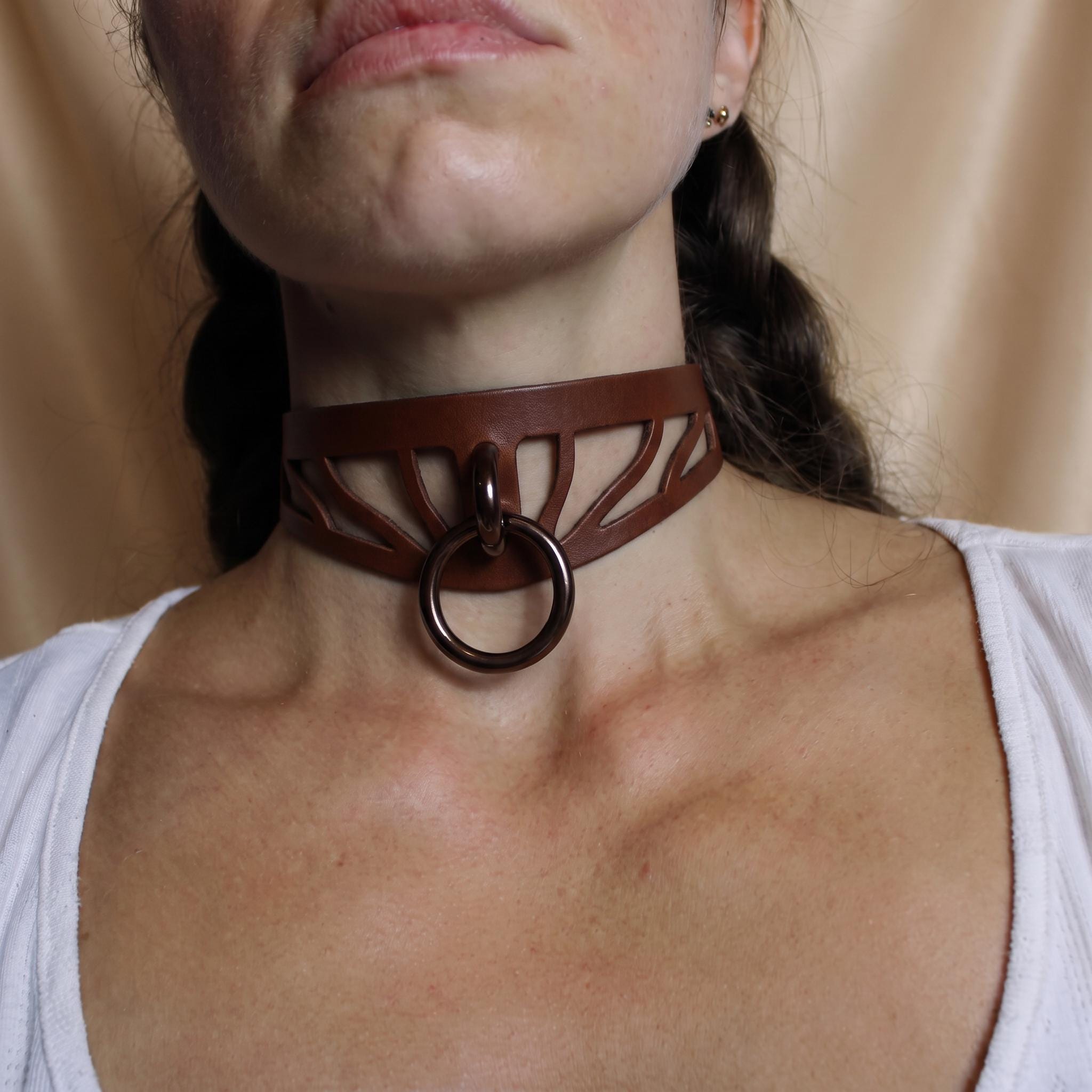 A close-up of a woman's neck and chest area wearing a brown leather choker necklace with a circular ring.