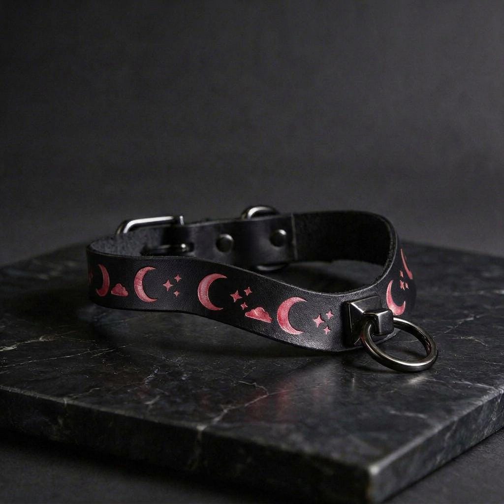 A black leather collar with a moon and star pattern design, featuring a metal ring and buckle.