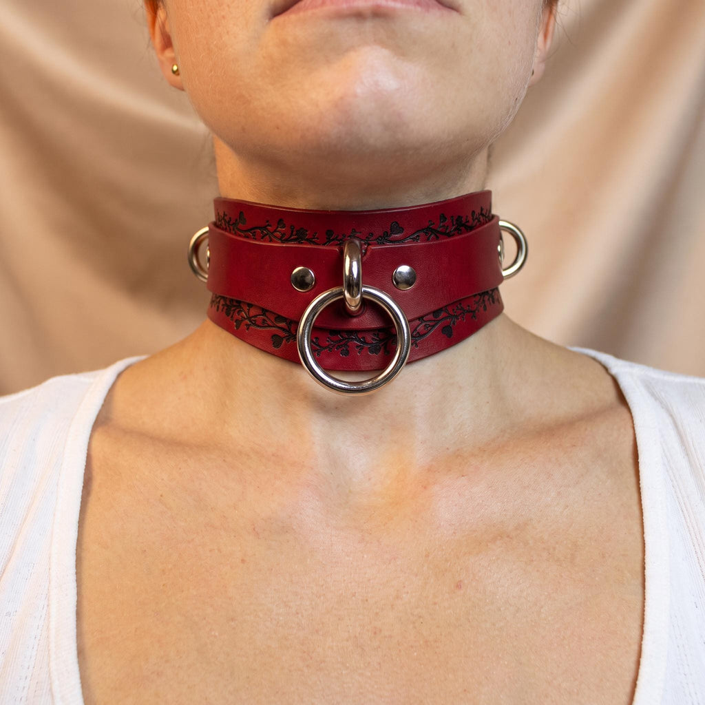 A close-up of a person wearing a red leather choker necklace with metal rings and a metal ring on the front.