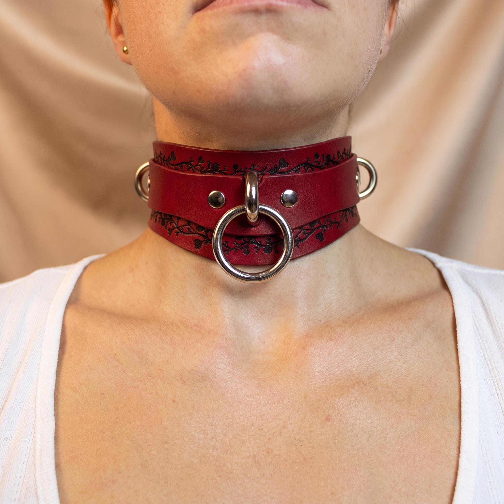 A close-up of a person wearing a red leather choker necklace with metal rings and a metal ring on the front.