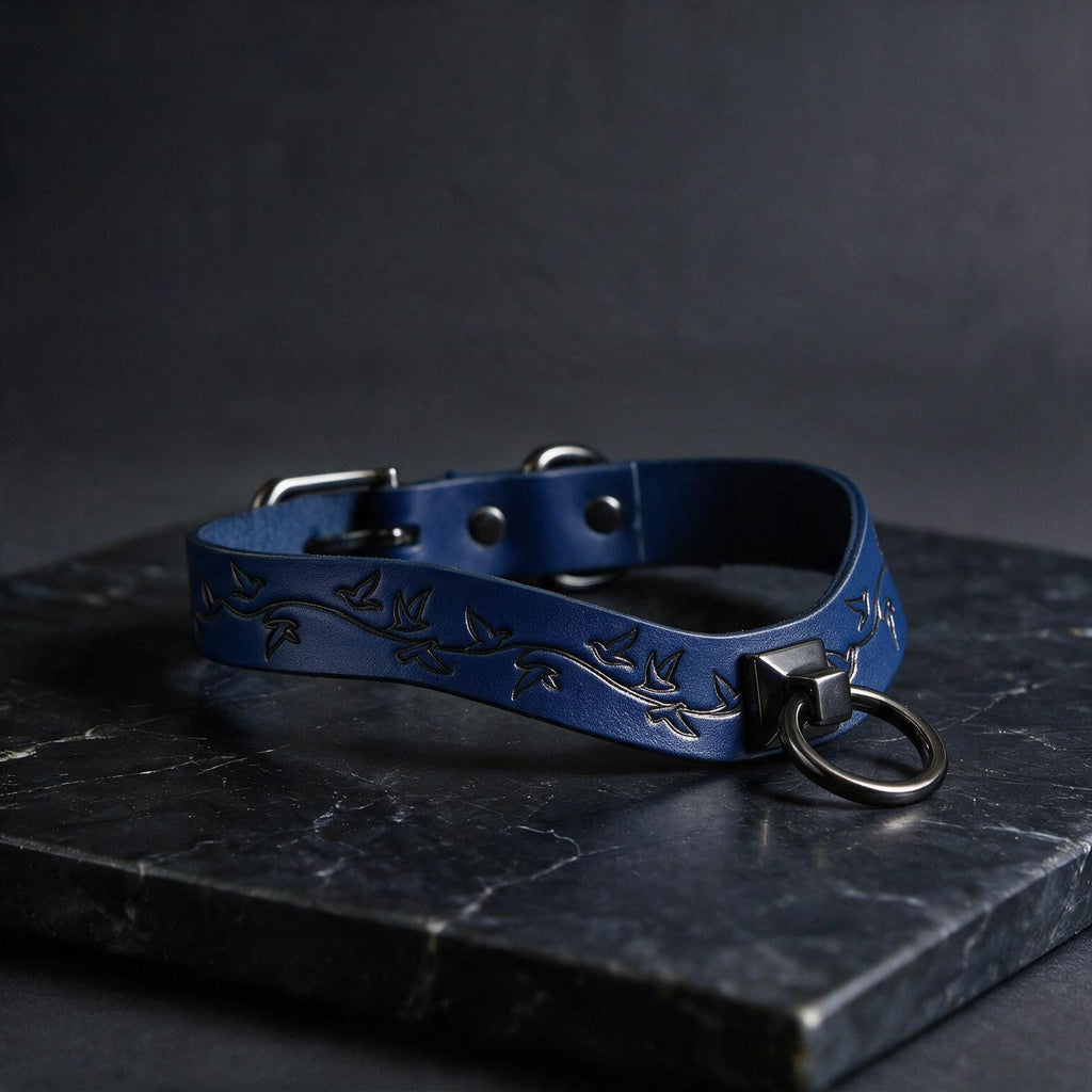 A blue leather collar with a metal ring and a leaf design, resting on a dark surface.