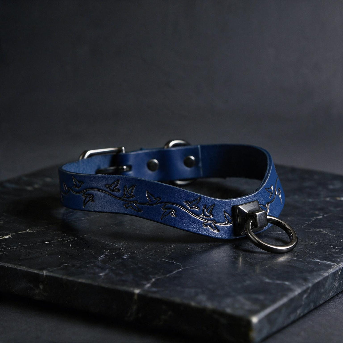 A blue leather collar with a metal ring and a leaf design, resting on a dark surface.