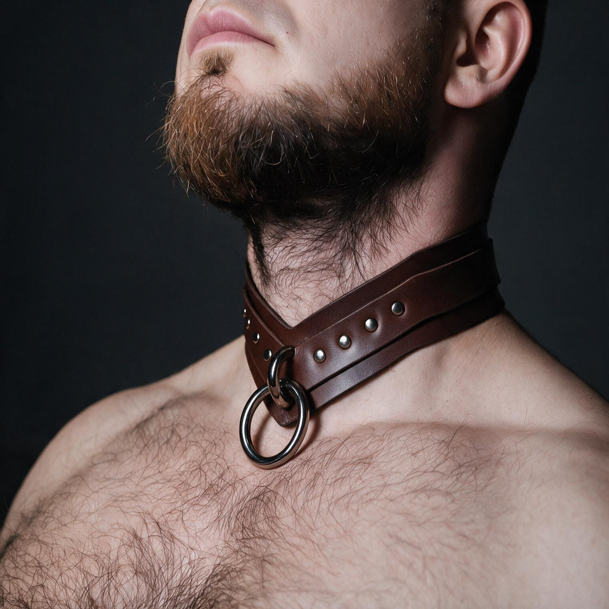 A close-up of a man's neck and chest area, with a leather choker collar featuring a metal ring and studs.