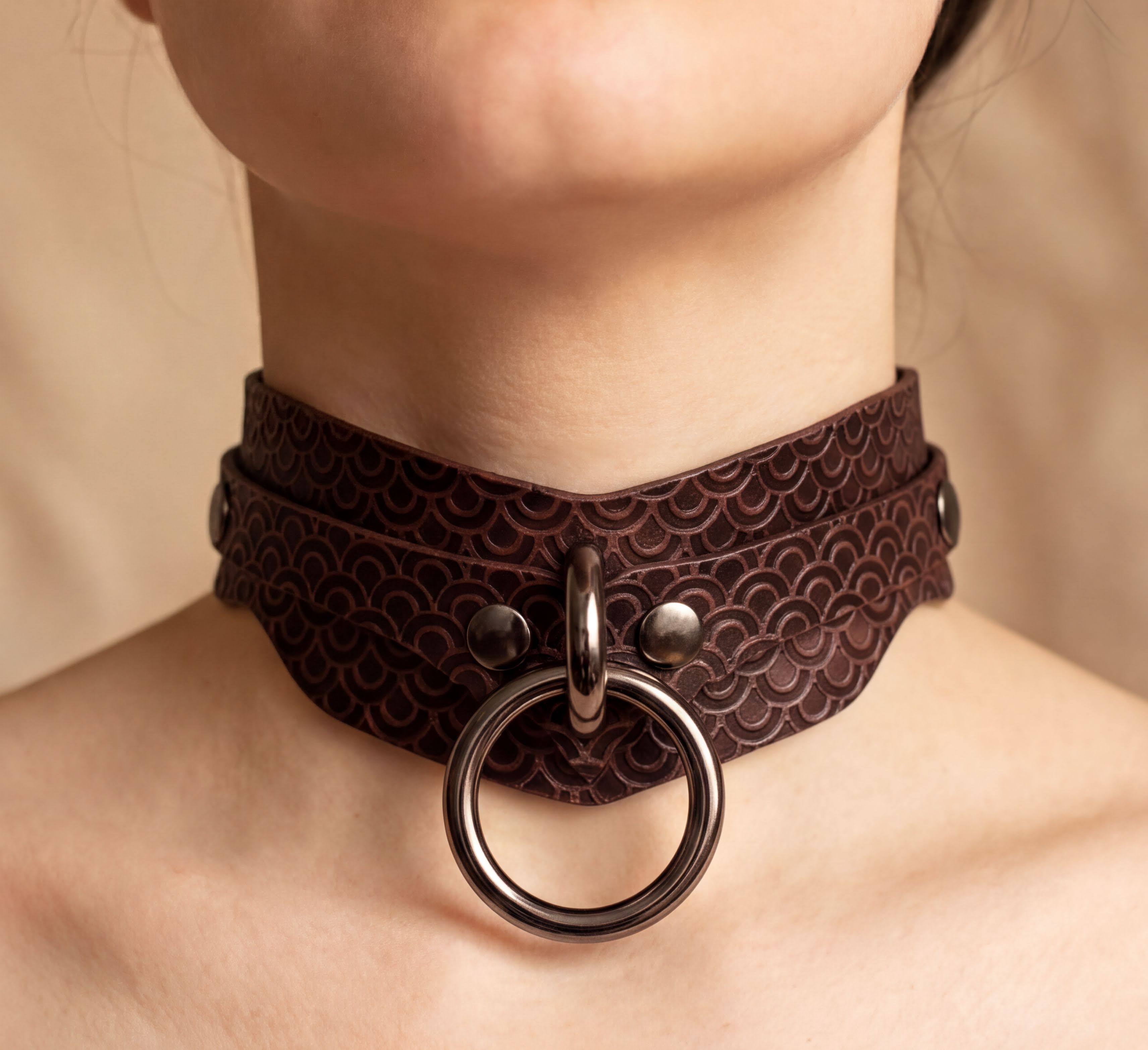A close-up of a woman's neck wearing a brown leather choker necklace with a metal ring and buckle.
