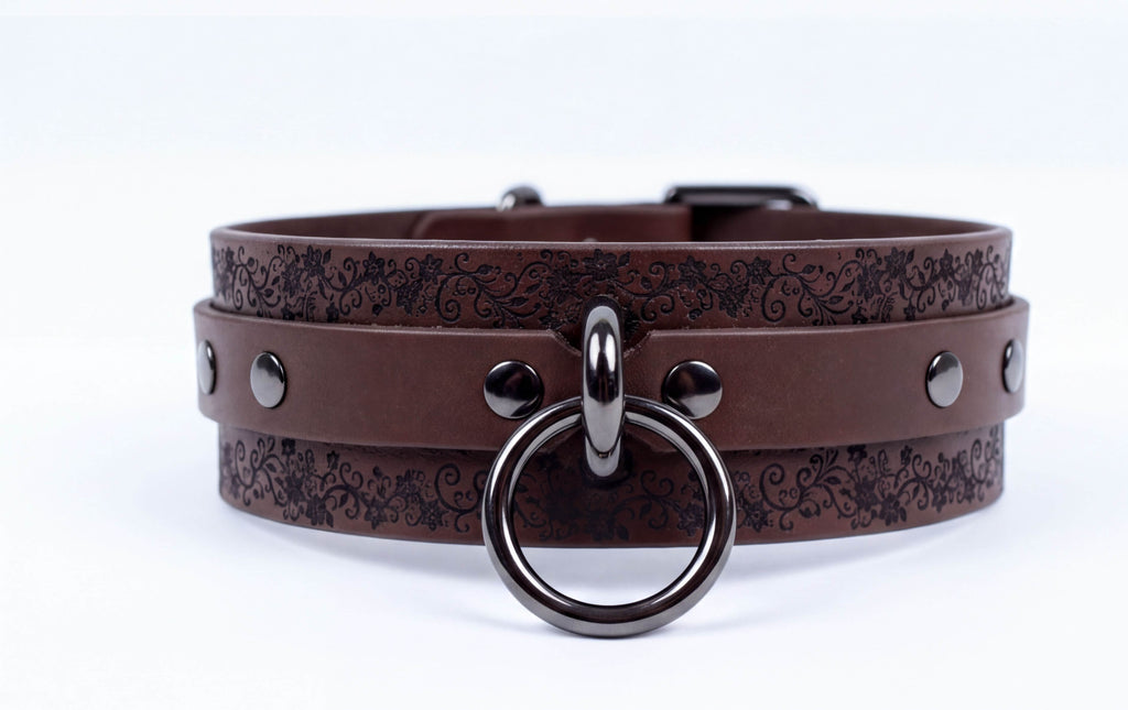 A brown leather choker collar with a metal ring and studs, featuring a floral pattern on the straps.