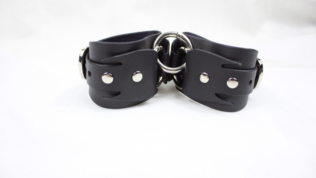 A pair of black leather cuffs with metal buckles and rivets, arranged in a symmetrical manner.