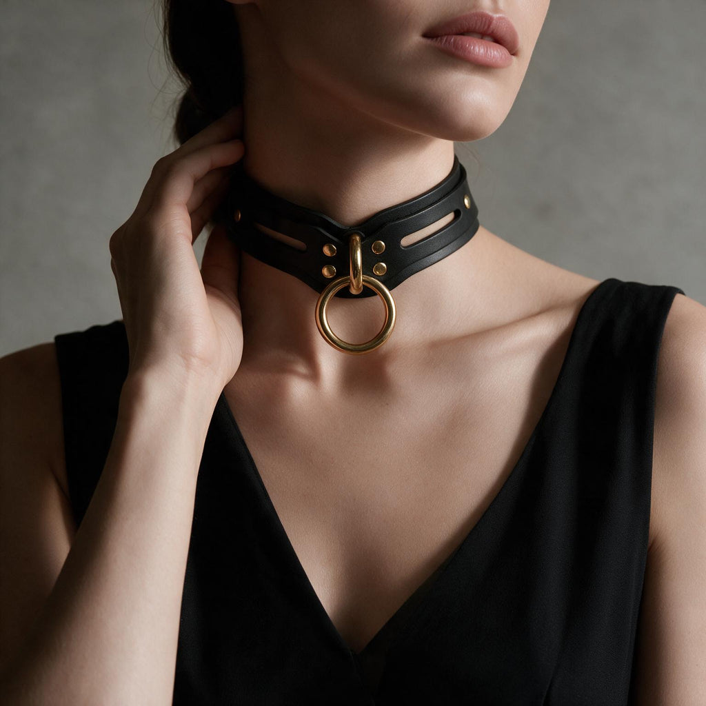 A close-up of a woman wearing a black choker necklace with a gold ring, posing against a gray background.