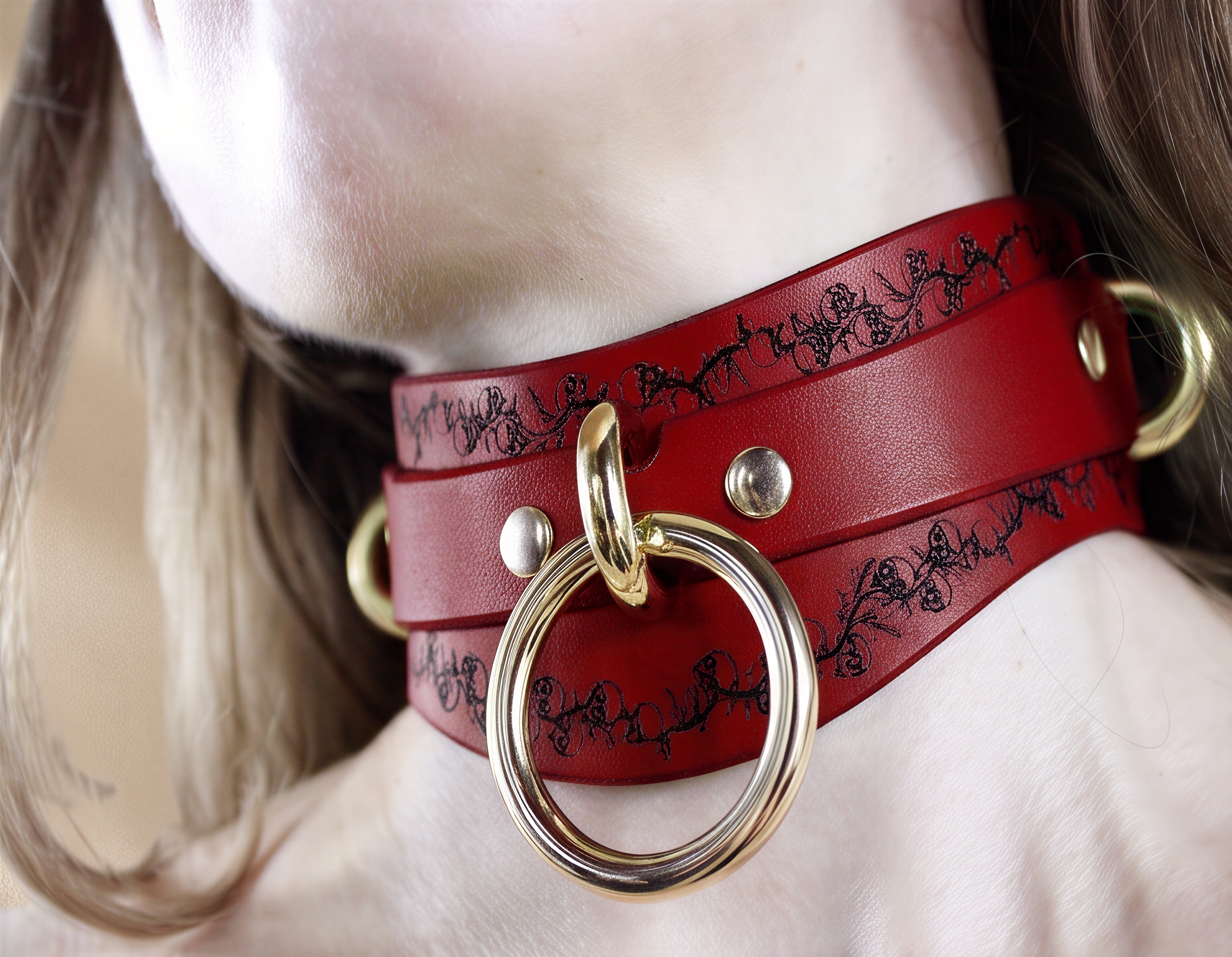 A close-up of a person's neck wearing a red leather choker with a gold ring and a black floral pattern.