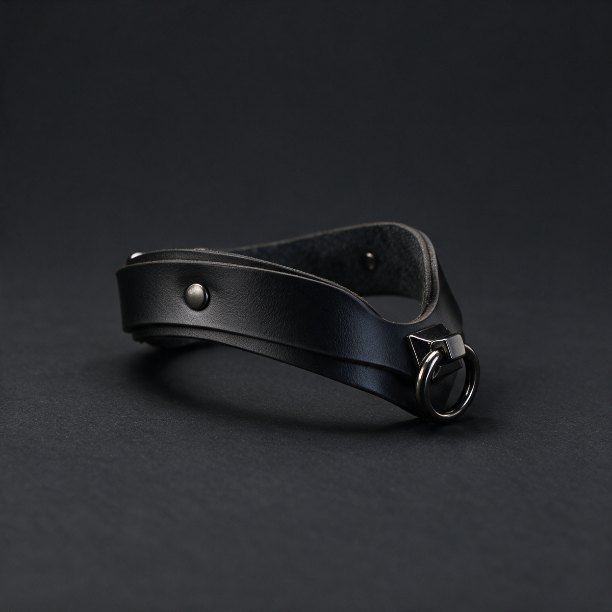 A black leather choker necklace with a metal clasp and a round metal ring is shown against a dark background.