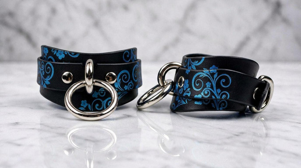 a pair of black leather straps with blue floral designs and silver metal rings, placed on a marble surface.