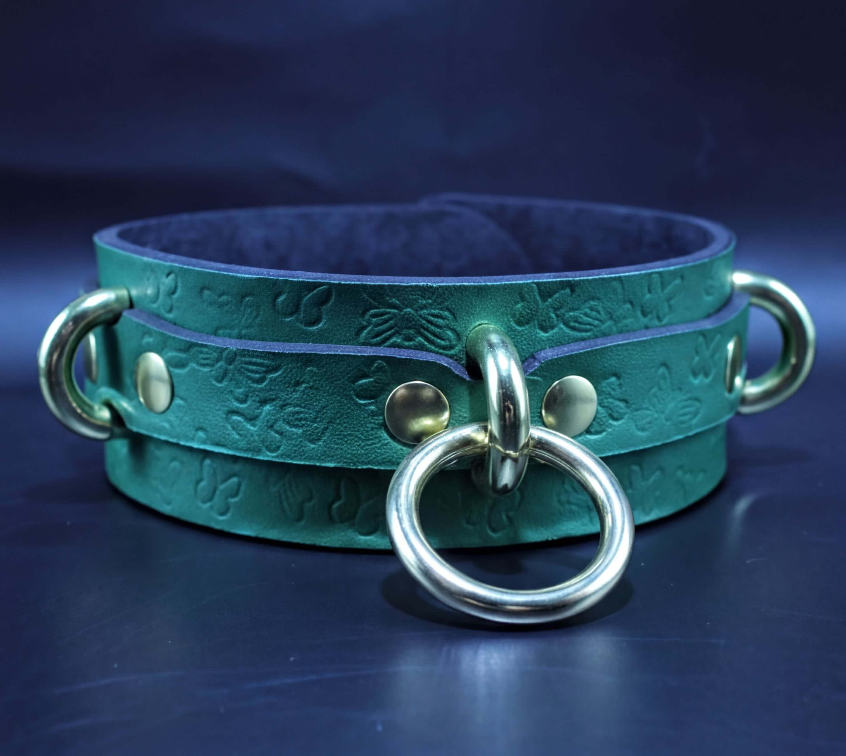 A green leather choker with a metal ring and two metal rings on either side, set against a dark blue background.
