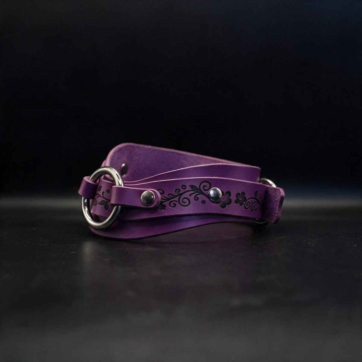 A purple leather belt with a metal buckle and floral design on the strap is shown against a dark background.