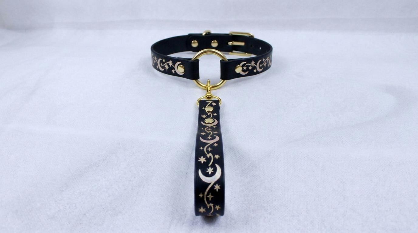 A black collar with a gold-colored buckle and a decorative strap featuring a moon and star pattern.