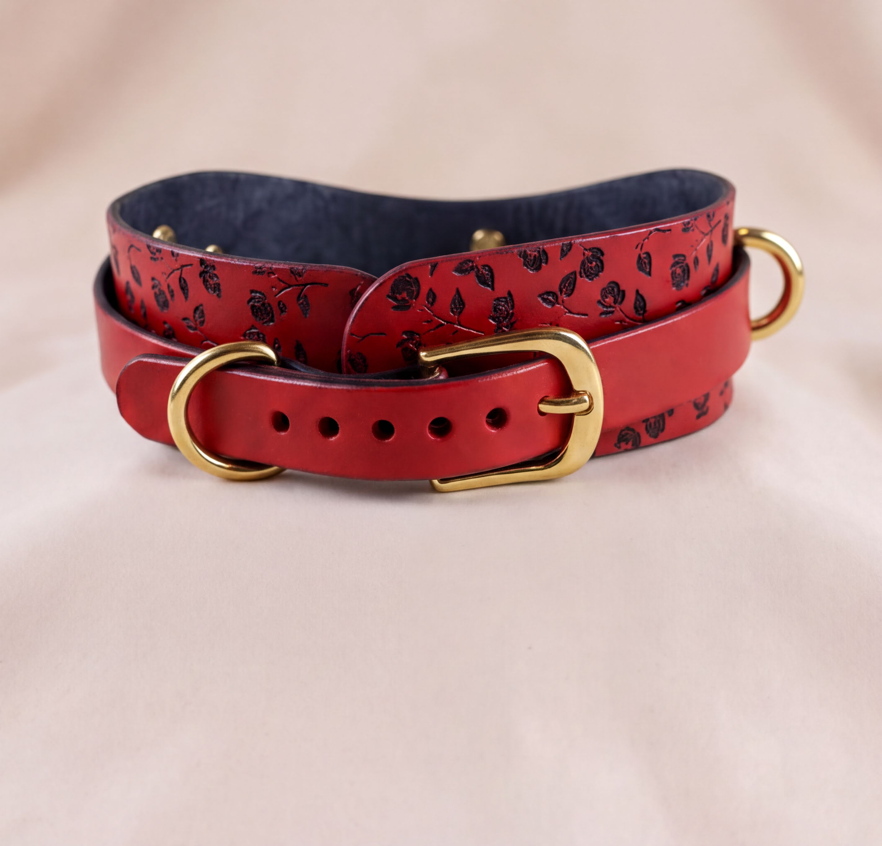A red leather belt with a floral pattern and gold hardware, including a buckle and two buckles, is displayed on a light-colored fabric background.