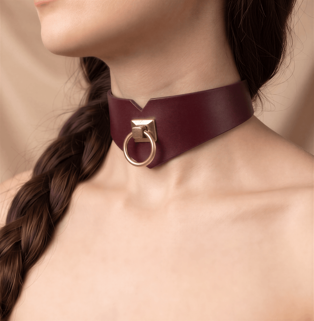 A close-up of a woman's neck and chest area wearing a burgundy leather choker necklace with a gold-colored metal ring.