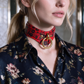 A close-up of a woman wearing a red choker necklace with a floral pattern and a gold ring on the front. She is wearing a blue floral blouse.
