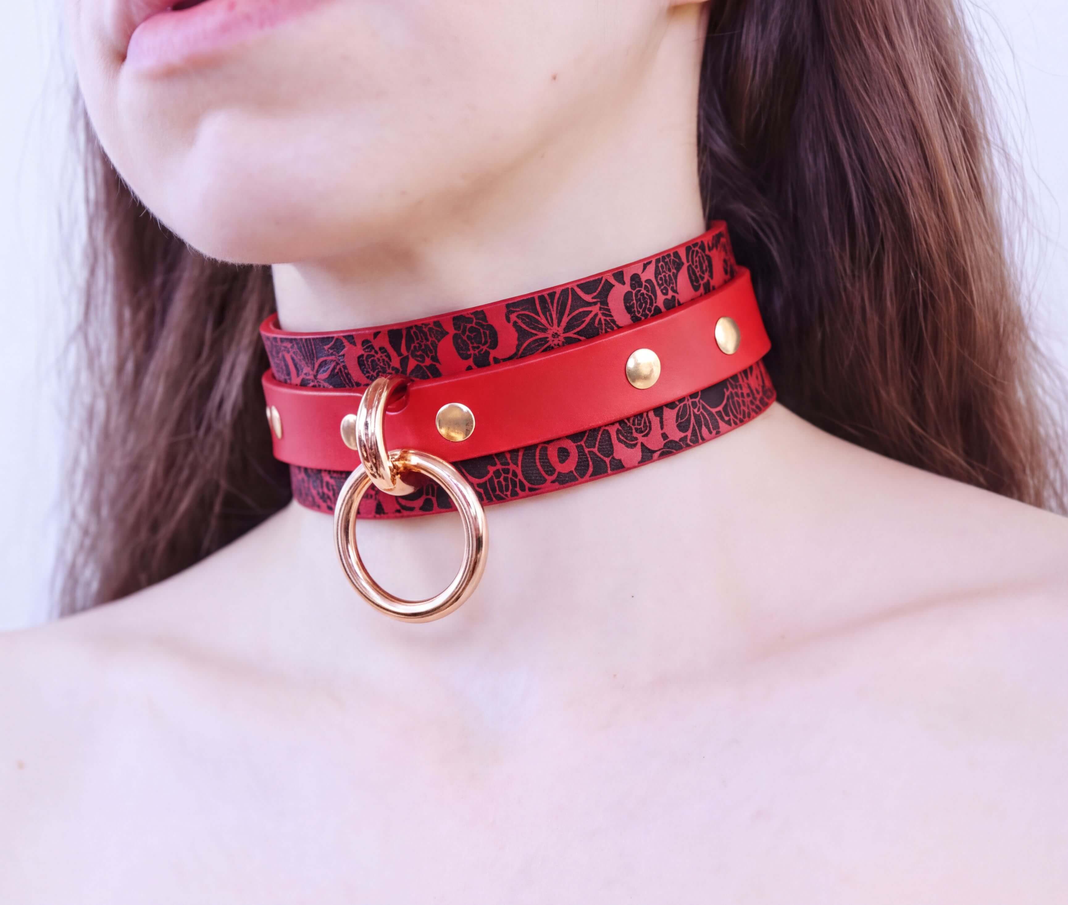 A close-up of a person's neck wearing a red choker necklace with a gold ring and a black floral pattern.
