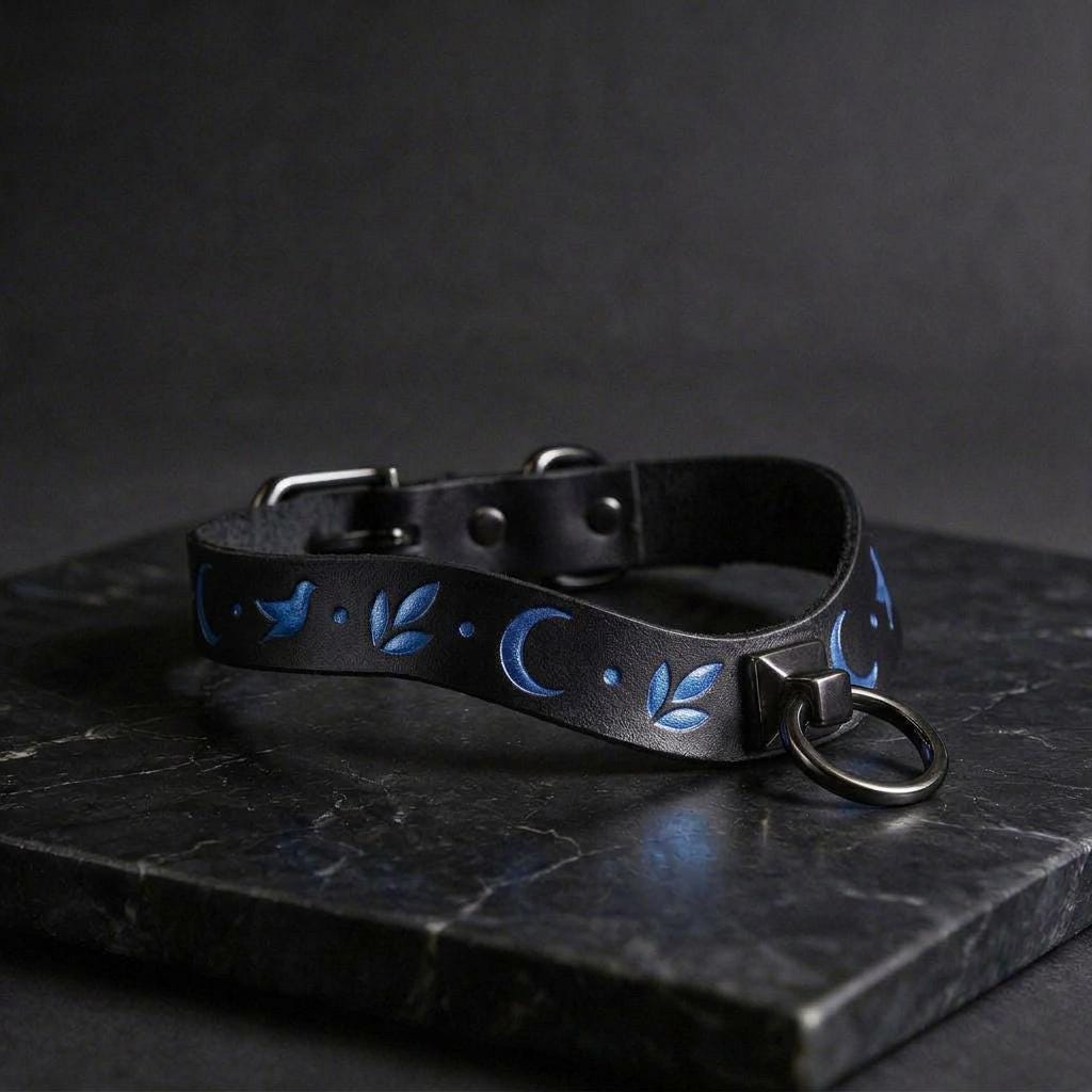 A black leather collar with blue floral designs and a metal ring on it, resting on a dark surface.