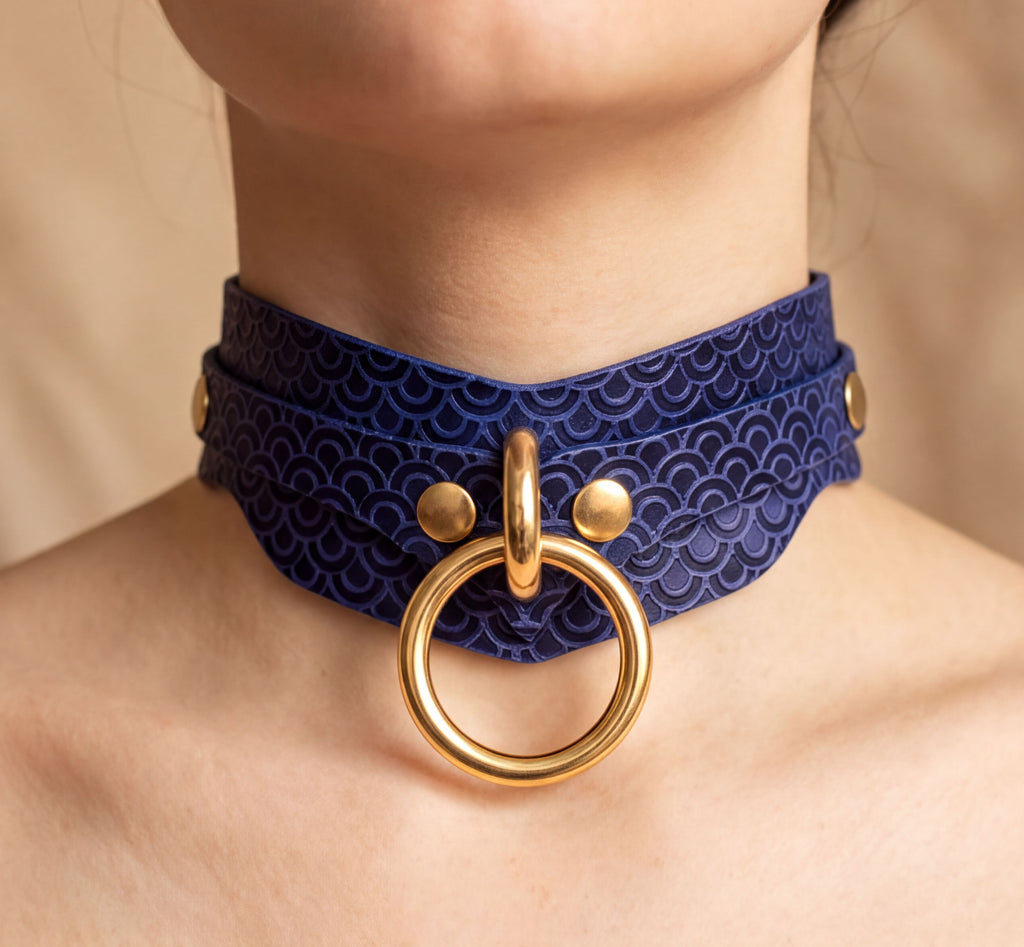 A close-up of a person's neck wearing a blue choker necklace with a gold ring and two gold-colored buttons.