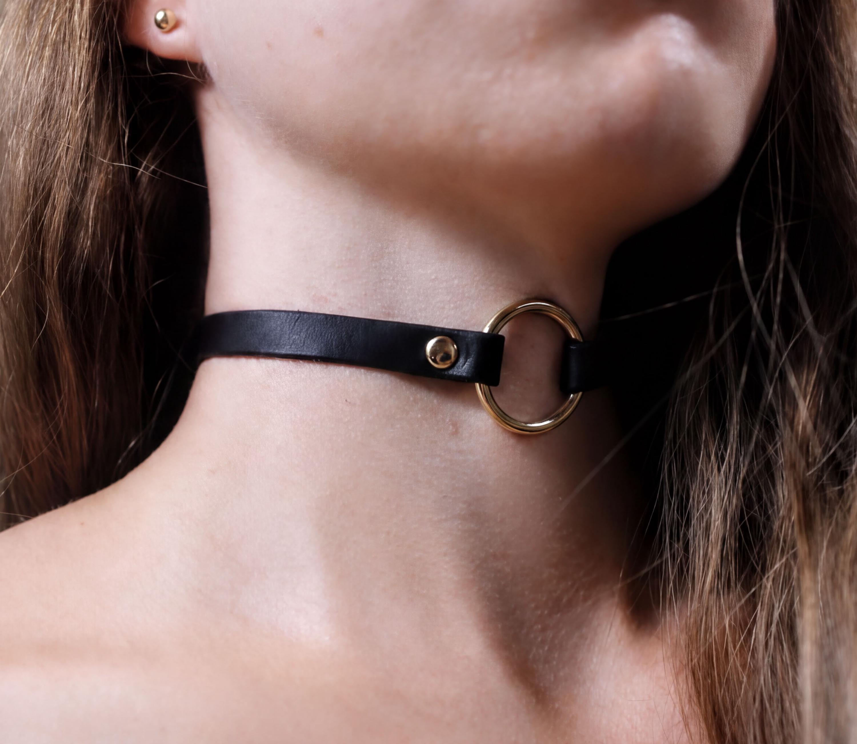 A close-up of a person's neck and chin wearing a black leather choker necklace with a gold ring.