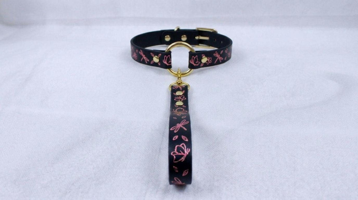 A black collar with a gold-colored buckle and a pink floral pattern on the strap is displayed on a white surface.