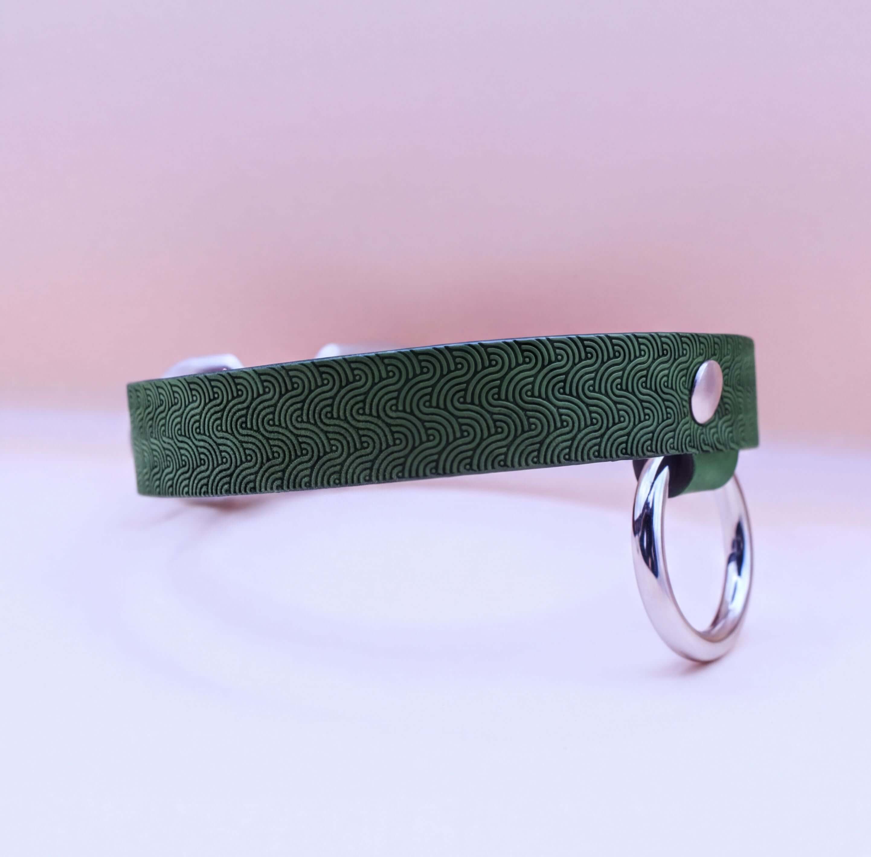 A green patterned collar with a silver ring attached, resting on a pink background.