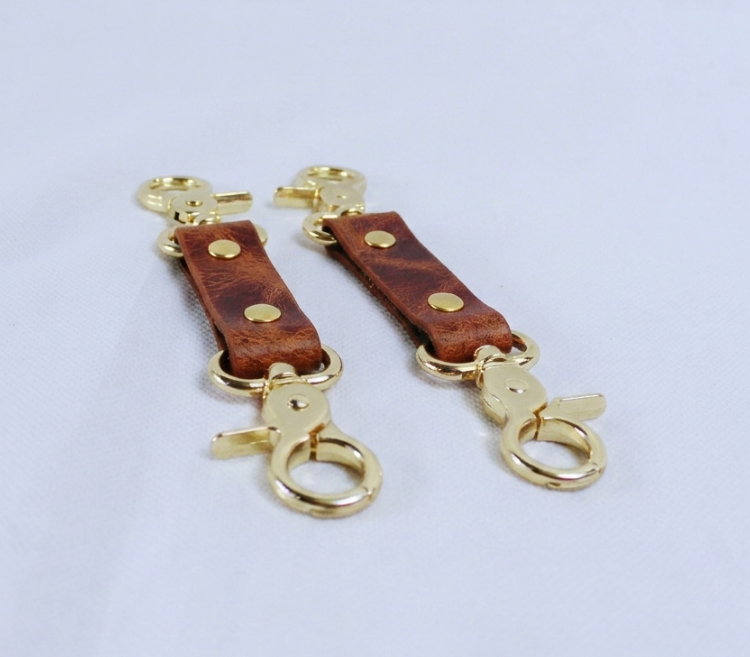 A pair of gold-colored leather keychains with metal clasps and loops, resting on a white surface.