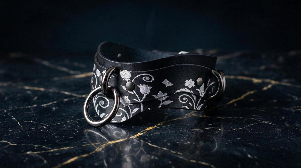 A black leather choker necklace with a floral pattern and metal rings on the front, resting on a dark marble surface.