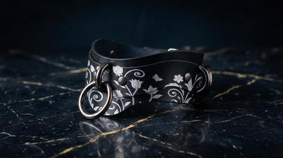 A black leather choker necklace with a floral pattern and metal rings on the front, resting on a dark marble surface.