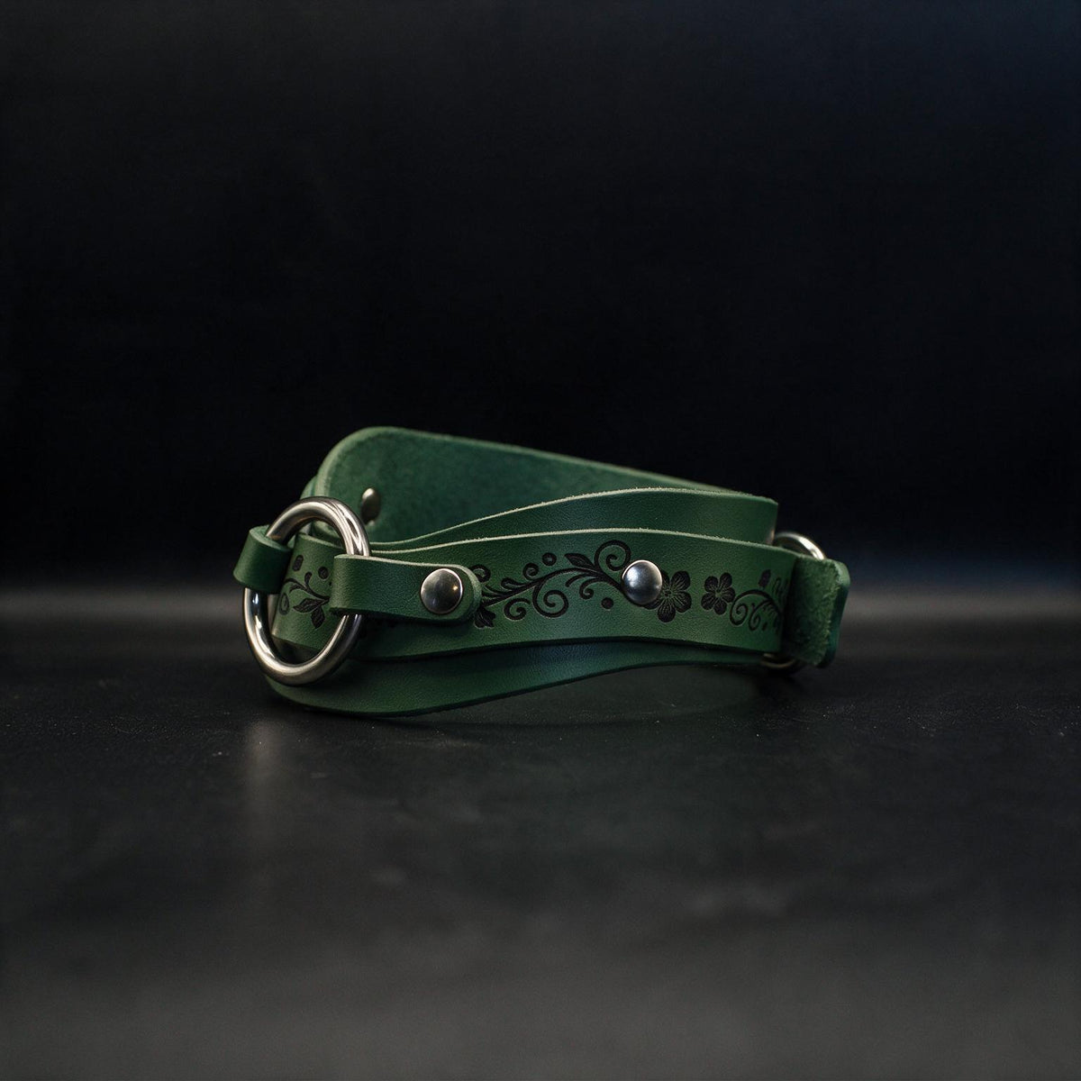 A green leather belt with a metal buckle and floral design on the strap is shown against a dark background.