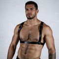 A shirtless man with a beard wearing a leather harness stands against a plain white background.