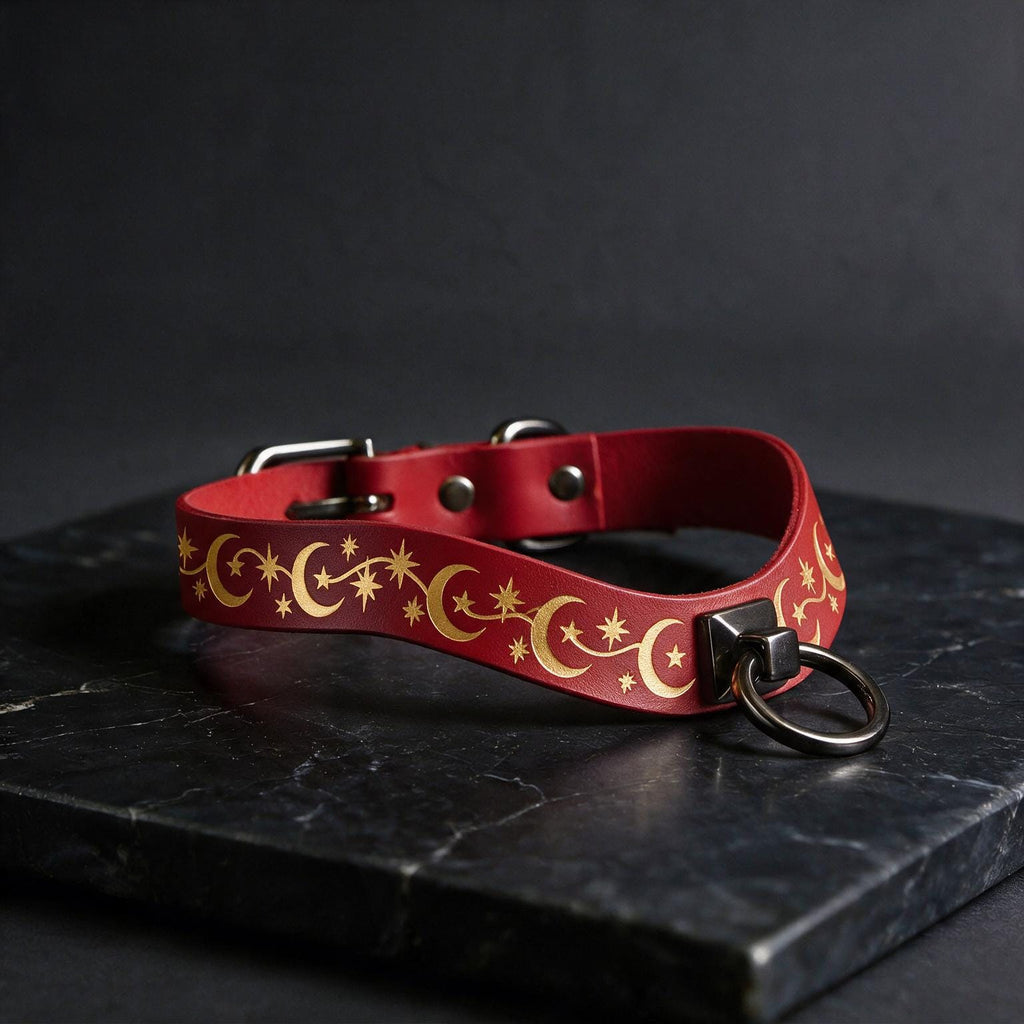 A red leather collar with a gold star and crescent design, featuring a metal ring and buckle, is displayed on a black marble surface.