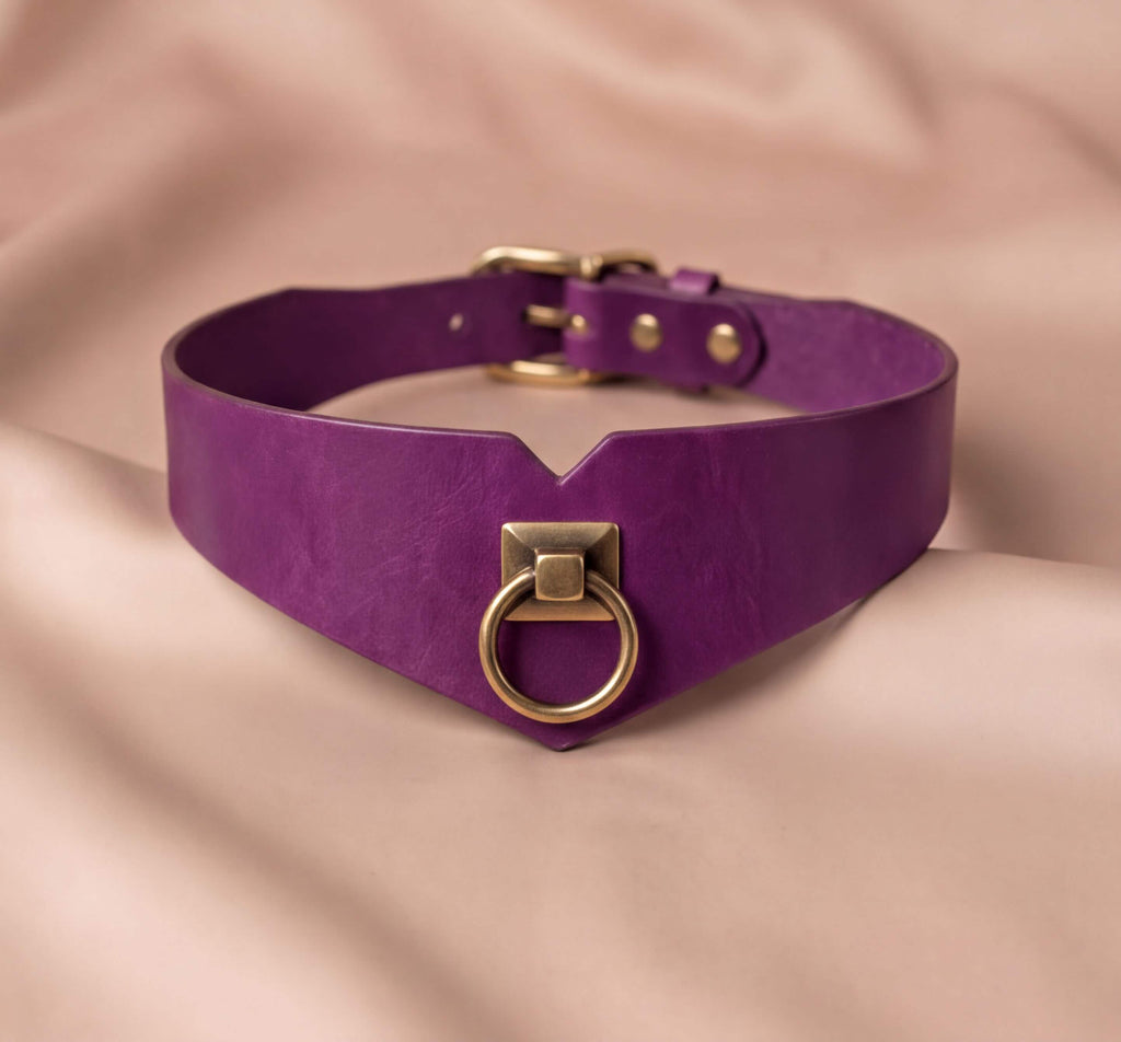 A purple choker collar with a gold ring and buckle is displayed on a beige fabric background.