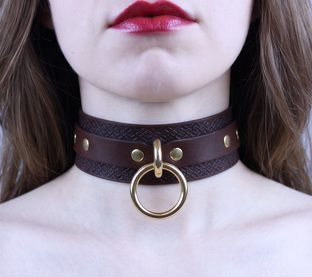 A close-up of a woman's neck and chest area, featuring a dark brown leather choker necklace with a gold ring and studs.