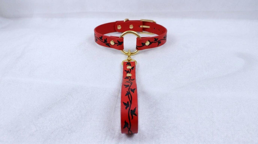 A red collar with a gold buckle and a black leaf design is displayed against a white background.