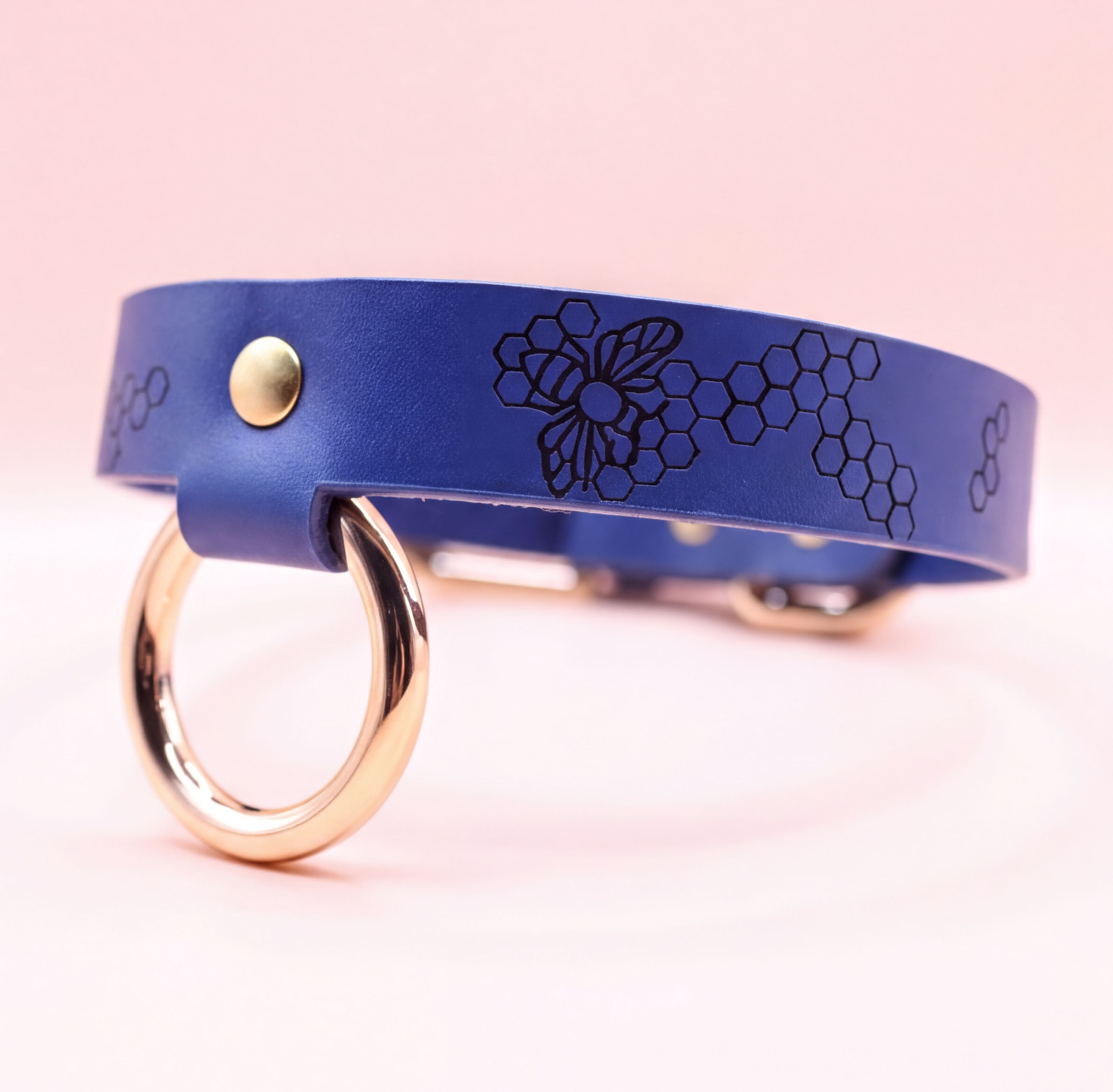 A blue leather collar with a gold-colored ring and a bee design printed on it.