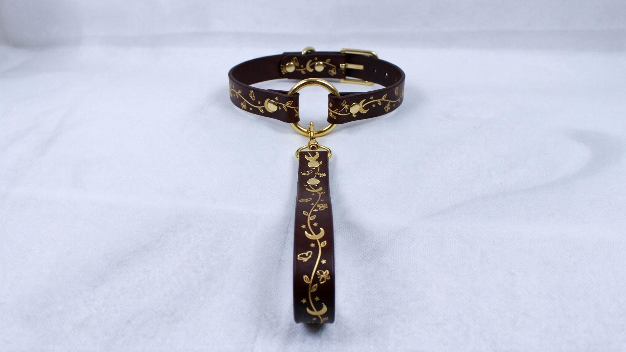 A brown leather collar with a gold-colored metal clasp and a decorative handle attached to it, resting on a white surface.