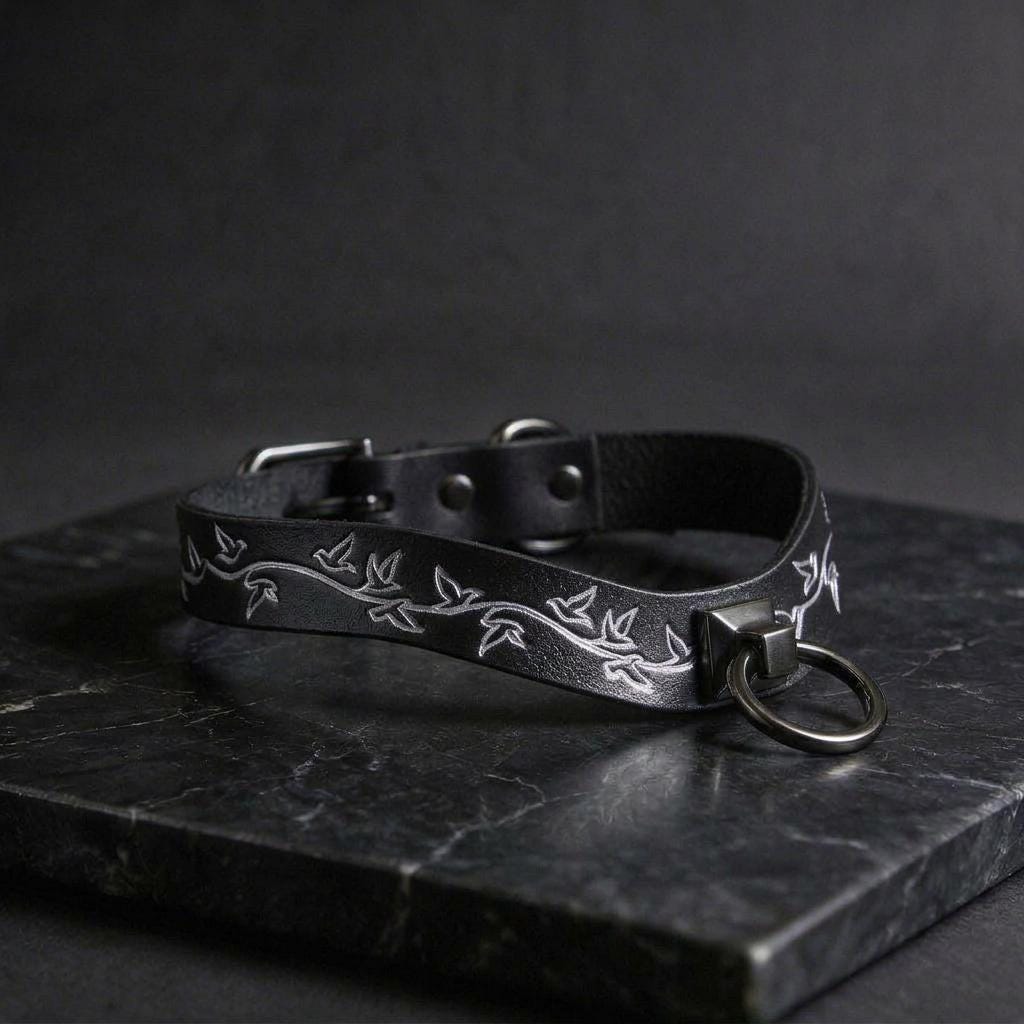 A black leather collar with a silver metal ring and a leaf design on it, resting on a dark surface.