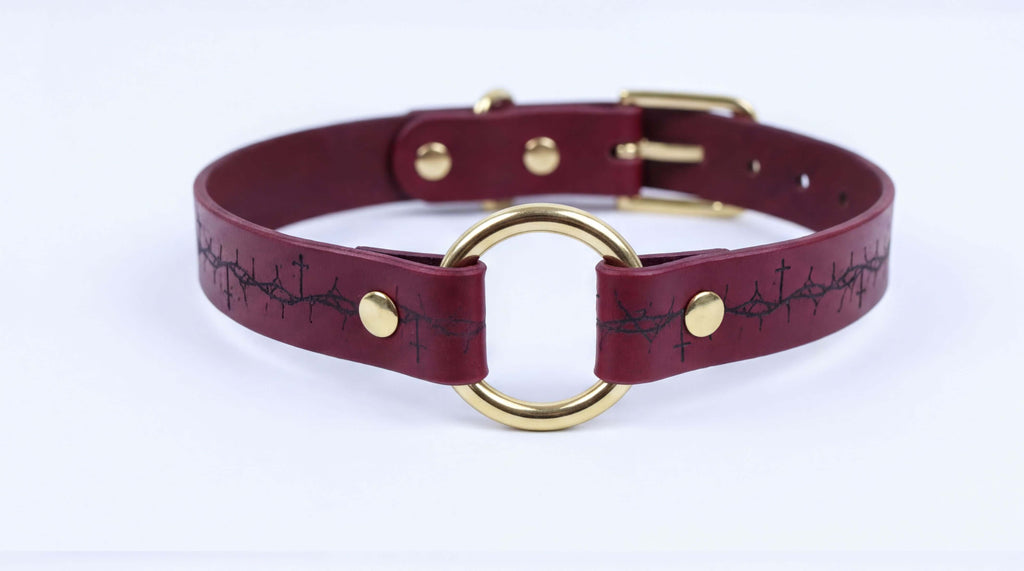 A burgundy leather choker collar with a gold-colored buckle and a circular ring.