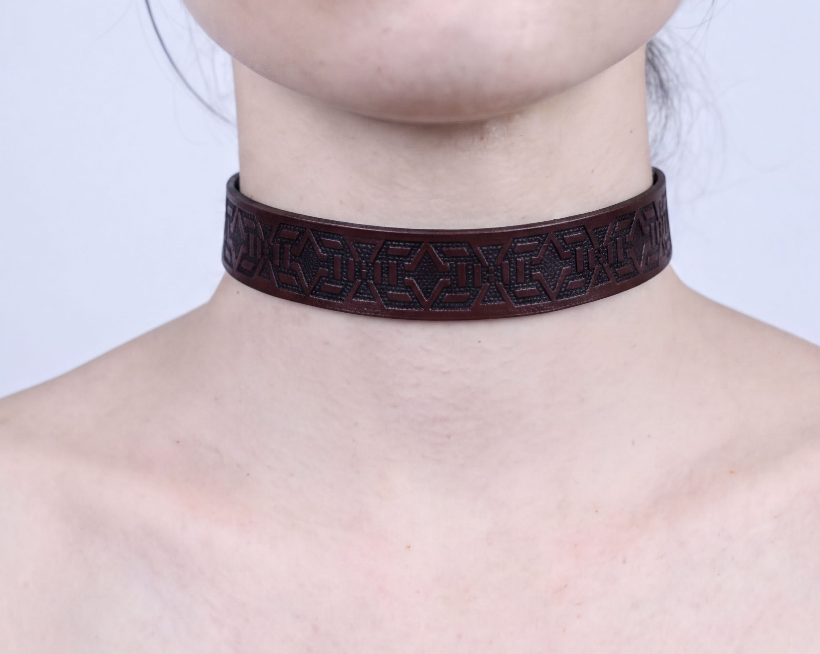 A close-up of a person's neck wearing a dark brown leather choker necklace with an intricate geometric pattern.