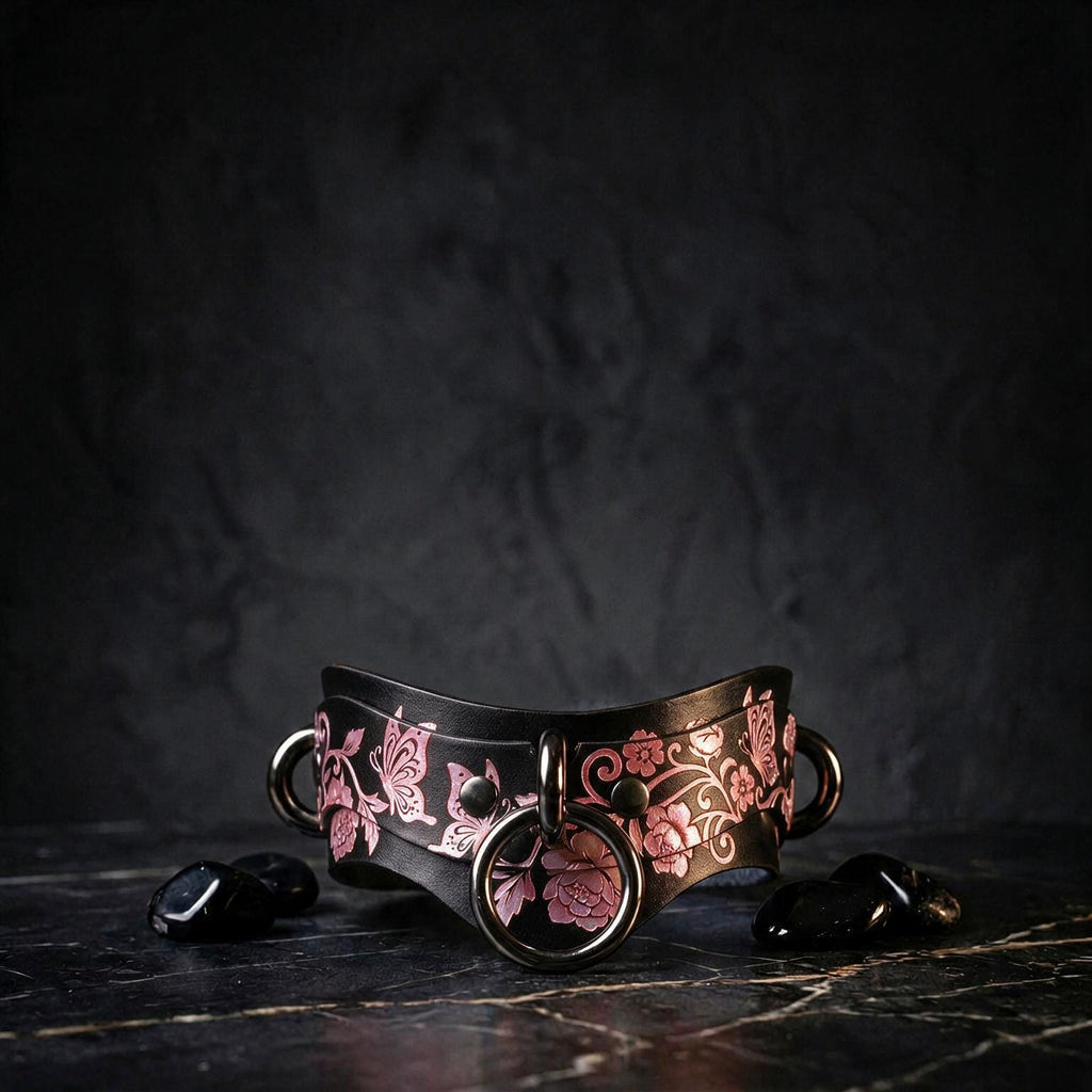 A black leather choker necklace with a floral design and metal rings is displayed on a dark surface, accompanied by small black stones.