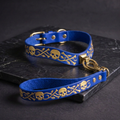 A blue leather dog collar with gold skull and bone designs, and a matching blue leash with a gold hook attached, both resting on a black surface.