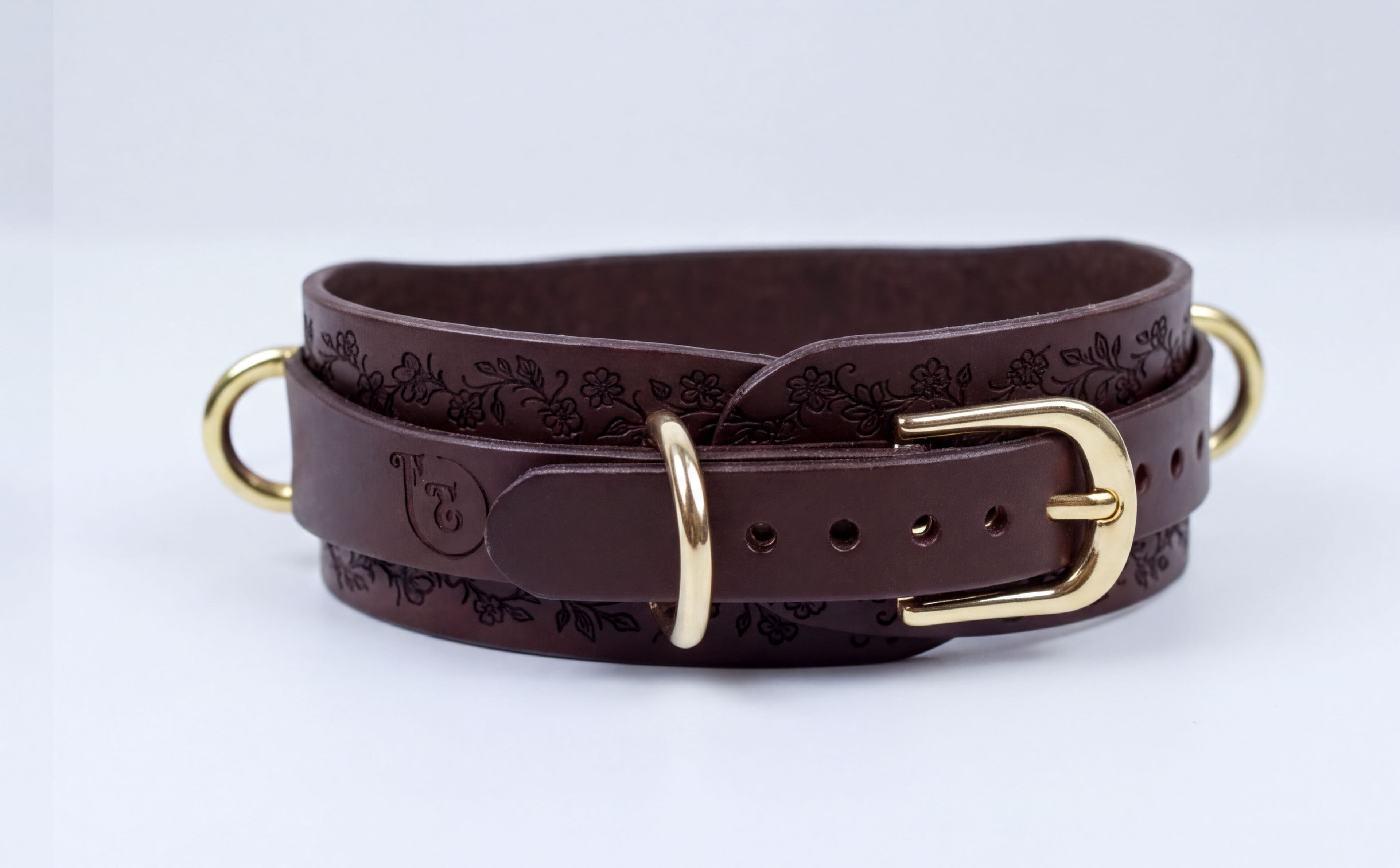 A brown leather belt with a gold buckle, featuring a floral pattern on the buckle and a round logo on the belt.
