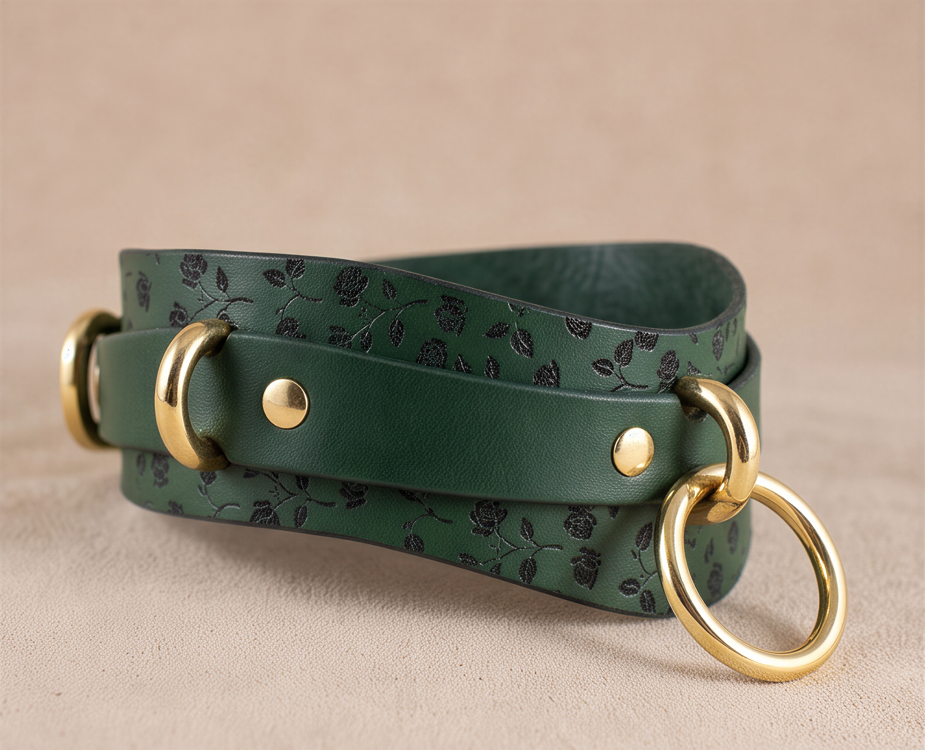 A green leather strap with a floral pattern and two gold-colored metal rings on the ends, resting on a beige surface.